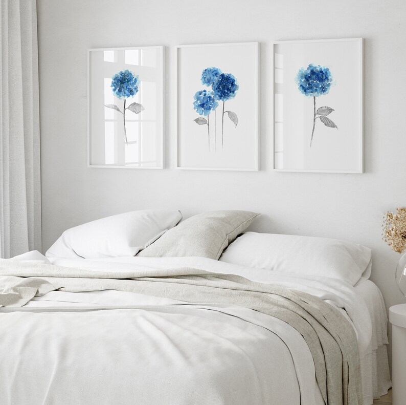 May include: Three watercolor paintings of blue hydrangeas on white backgrounds, displayed in a row on a white wall above a white bed with a light gray blanket.