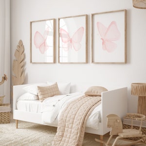May include: Three framed watercolor paintings of pink butterflies on a white wall above a white daybed with a beige quilt and pillows.