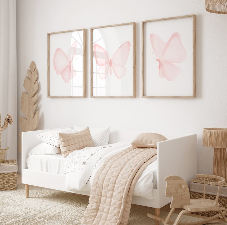 May include: Three framed watercolor paintings of pink butterflies on a white wall above a white daybed with a beige quilt and pillows.