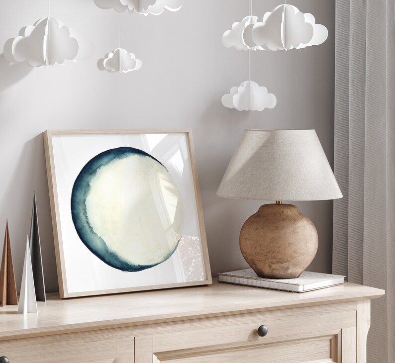 K&ouml;nnte beinhalten: Ein gerahmtes Aquarellbild eines blauen und wei&szlig;en Kreises auf wei&szlig;em Hintergrund. Das Bild h&auml;ngt an einer hellbraunen Wand mit einer Lampe und einem Buch auf einem Tisch darunter.