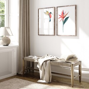 May include: A wooden bench with a white throw blanket and a book on top. Two framed watercolor prints hang on the wall behind the bench. One print features a hummingbird in flight, the other a red flower.