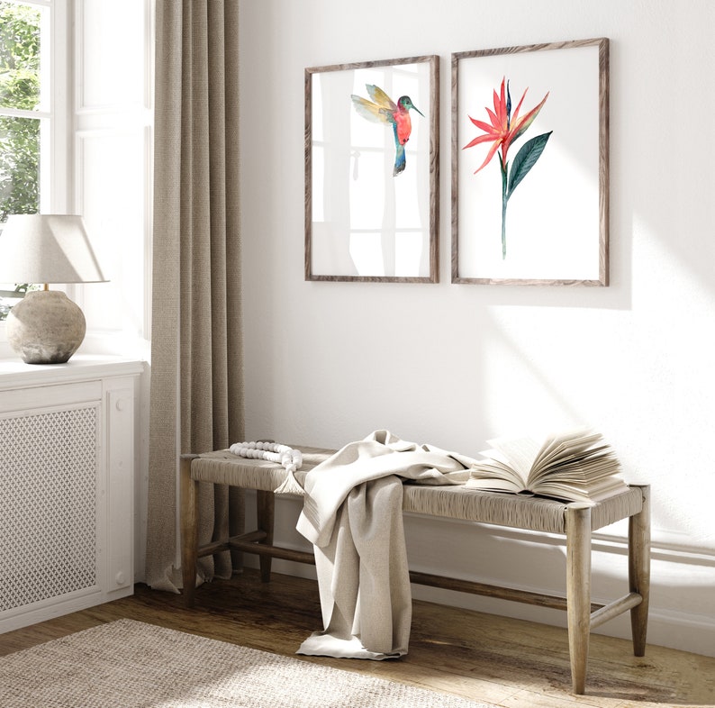 May include: A wooden bench with a white throw blanket and a book on top. Two framed watercolor prints hang on the wall behind the bench. One print features a hummingbird in flight, the other a red flower.