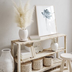 K&ouml;nnte beinhalten: Ein zweist&ouml;ckiges Holzregal mit einer wei&szlig;en Vase mit getrocknetem Pampasgras, einem gerahmten Aquarelldruck einer blauen Blume, einem Stapel B&uuml;cher, einer wei&szlig;en Vase mit dekorativem Design und drei geflochtenen K&ouml;rben.