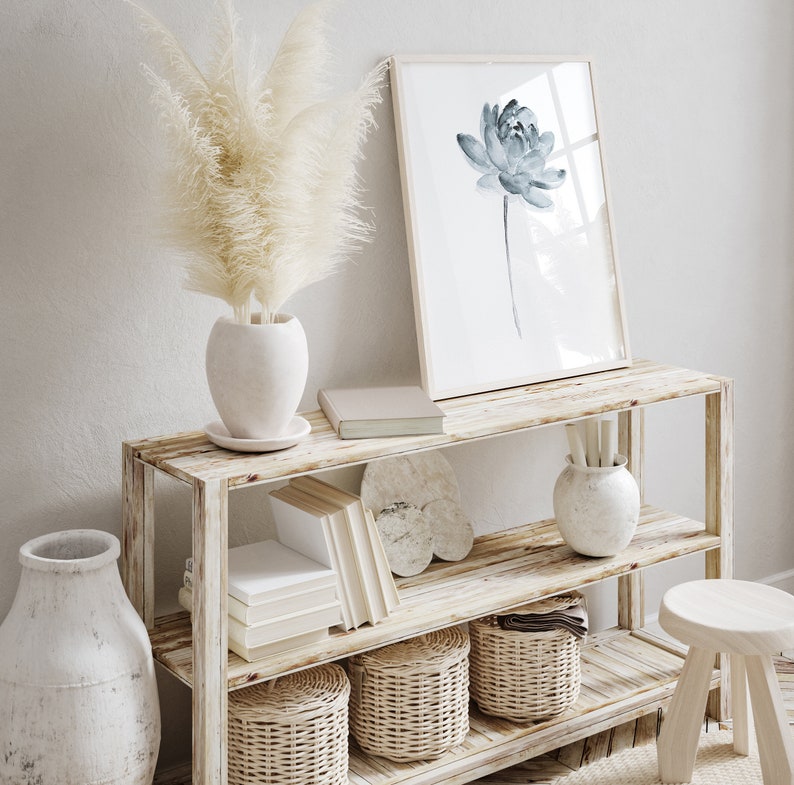 K&ouml;nnte beinhalten: Ein zweist&ouml;ckiges Holzregal mit einer wei&szlig;en Vase mit getrocknetem Pampasgras, einem gerahmten Aquarelldruck einer blauen Blume, einem Stapel B&uuml;cher, einer wei&szlig;en Vase mit dekorativem Design und drei geflochtenen K&ouml;rben.