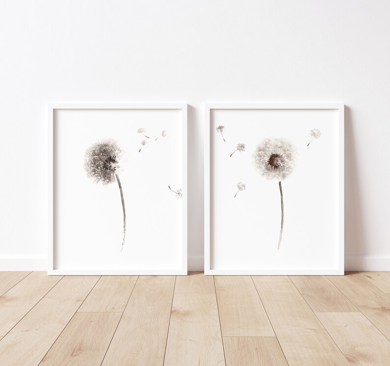 May include: Two framed black and white watercolor paintings of dandelions with seeds blowing in the wind. The paintings are on a light wood floor.