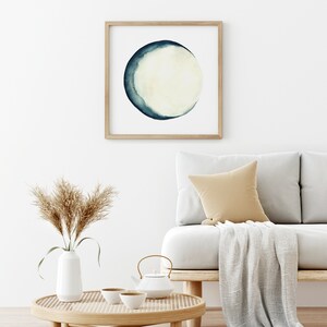 K&ouml;nnte beinhalten: Ein gerahmtes Aquarellbild eines Halbmondes in Blaut&ouml;nen und Wei&szlig;, das an einer wei&szlig;en Wand &uuml;ber einem hellgrauen Sofa mit einer beigefarbenen Wolldecke und einem braunen Kissen h&auml;ngt.