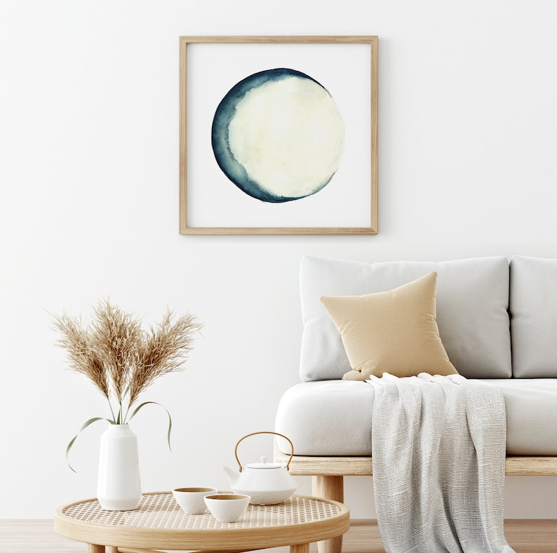 K&ouml;nnte beinhalten: Ein gerahmtes Aquarellbild eines Halbmondes in Blaut&ouml;nen und Wei&szlig;, das an einer wei&szlig;en Wand &uuml;ber einem hellgrauen Sofa mit einer beigefarbenen Wolldecke und einem braunen Kissen h&auml;ngt.