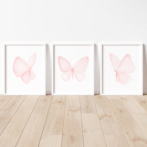 May include: Three white picture frames with pink watercolor butterfly illustrations inside. The frames are on a light wood floor.