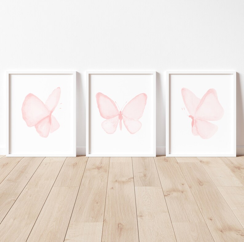 May include: Three white picture frames with pink watercolor butterfly illustrations inside. The frames are on a light wood floor.