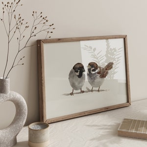 May include: A framed watercolor painting of two brown sparrows perched on a branch. The sparrows are facing each other and have black eyes. The background is a light brown with a watercolor wash of light blue and green.