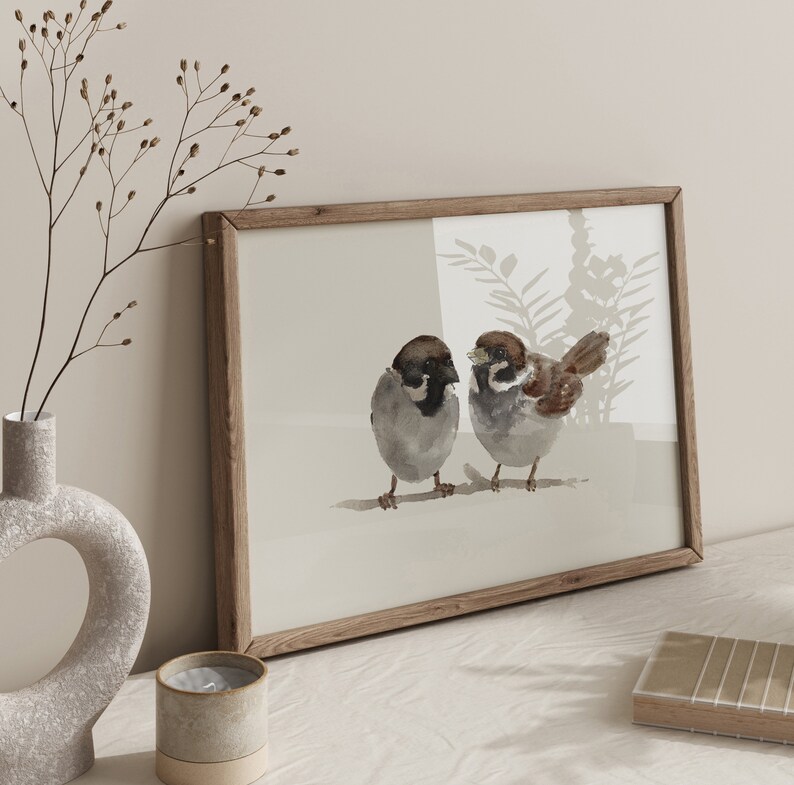 May include: A framed watercolor painting of two brown sparrows perched on a branch. The sparrows are facing each other and have black eyes. The background is a light brown with a watercolor wash of light blue and green.