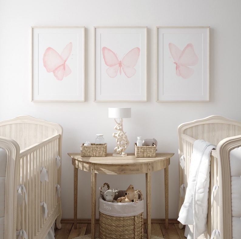 May include: Three framed watercolor paintings of pink butterflies on a white wall above two white cribs with white bedding. A wooden table with a lamp and a basket of toys sits between the cribs.