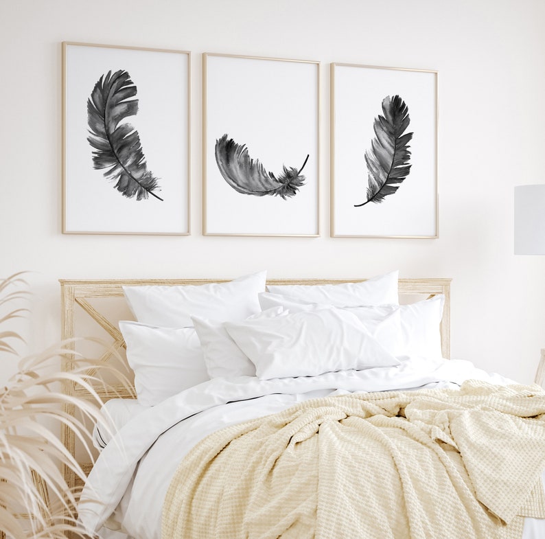 K&ouml;nnte beinhalten: Drei Schwarz-Wei&szlig;-Aquarell-Federdrucke in schlichten goldenen Rahmen, die an einer wei&szlig;en Wand &uuml;ber einem Bett mit wei&szlig;en Bettw&auml;sche und einer cremefarbenen Wolldecke h&auml;ngen.