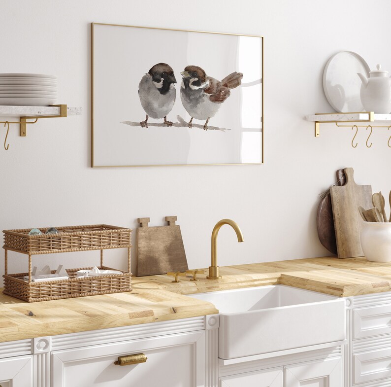 May include: A framed watercolor painting of two brown and gray sparrows perched on a branch. The painting is hanging on a white wall above a kitchen sink with a gold faucet.