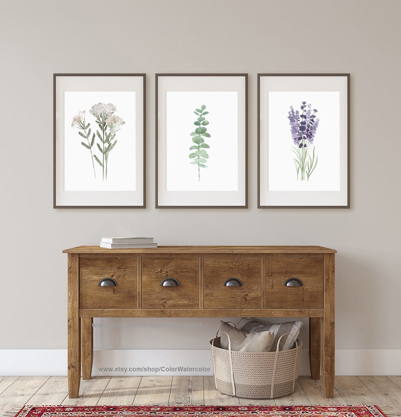 May include: Three framed watercolor prints of botanical illustrations. The prints feature a sprig of white flowers, a sprig of eucalyptus leaves, and a bunch of lavender flowers. The prints are displayed on a wall above a wooden console table.