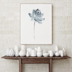 K&ouml;nnte beinhalten: Eine wei&szlig;e Backsteinwand mit einem gerahmten Aquarellbild einer blauen Blume. Ein Holztisch mit einer Auswahl an wei&szlig;en Keramikvasen und Schalen.