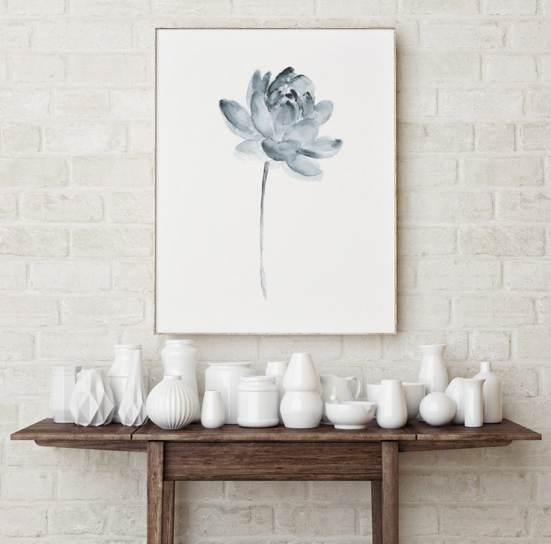 K&ouml;nnte beinhalten: Eine wei&szlig;e Backsteinwand mit einem gerahmten Aquarellbild einer blauen Blume. Ein Holztisch mit einer Auswahl an wei&szlig;en Keramikvasen und Schalen.