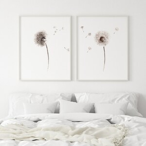 May include: Two framed watercolor paintings of dandelion flowers with seeds blowing in the wind. The paintings are in shades of brown and white and are set against a white wall.