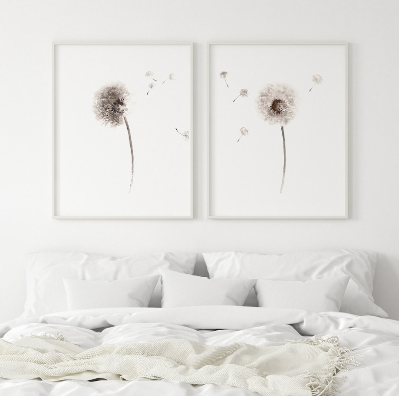 May include: Two framed watercolor paintings of dandelion flowers with seeds blowing in the wind. The paintings are in shades of brown and white and are set against a white wall.