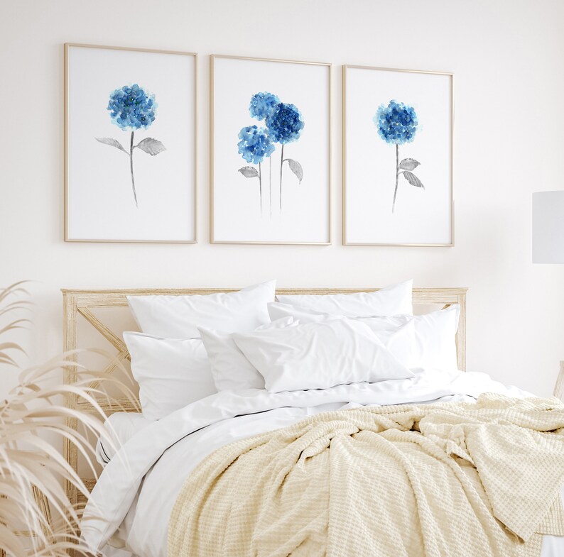May include: Three framed watercolor paintings of blue hydrangeas on a white wall above a bed with white bedding and a cream throw blanket.