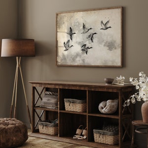 May include: A framed watercolor print of six ducks in flight, set against a muted, cloudy background. The artwork hangs above a rustic, dark wood storage cabinet with woven baskets, decorative items, and a pair of beige sandals. A floor lamp with a brown shade and a brown ottoman are also visible.