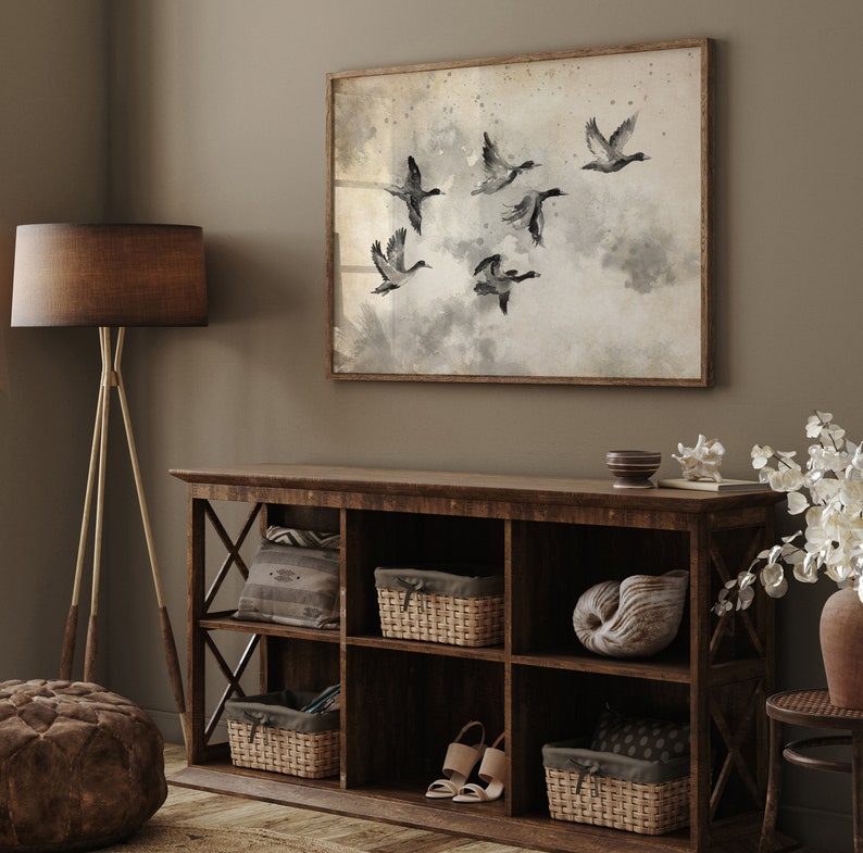 May include: A framed watercolor print of six ducks in flight, set against a muted, cloudy background. The artwork hangs above a rustic, dark wood storage cabinet with woven baskets, decorative items, and a pair of beige sandals. A floor lamp with a brown shade and a brown ottoman are also visible.