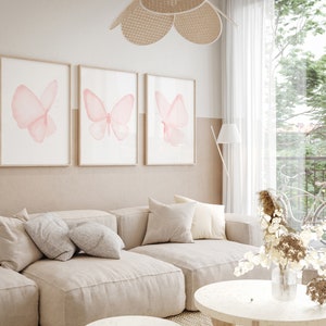 May include: Three framed prints of pink watercolor butterflies on a white wall above a beige sectional sofa with pillows. A vase of dried flowers sits on a round coffee table.