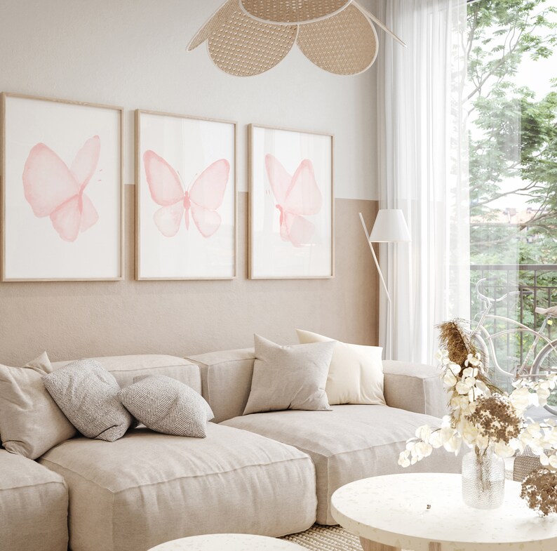 May include: Three framed prints of pink watercolor butterflies on a white wall above a beige sectional sofa with pillows. A vase of dried flowers sits on a round coffee table.