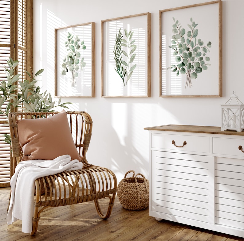 May include: A white cabinet with two drawers and two doors, a wicker chair with a brown cushion and a white throw blanket, and three framed watercolor prints of green leaves on a white wall.