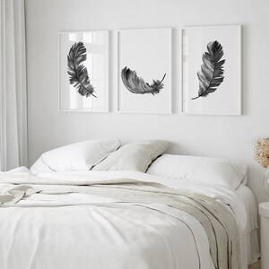 K&ouml;nnte beinhalten: Drei Schwarz-Wei&szlig;-Aquarell-Federdrucke in wei&szlig;en Rahmen an einer wei&szlig;en Wand &uuml;ber einem wei&szlig;en Bett mit einer wei&szlig;en Bettdecke und einer hellgrauen Tagesdecke.