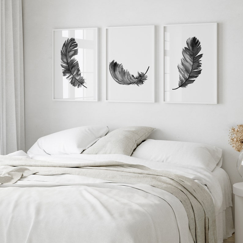 K&ouml;nnte beinhalten: Drei Schwarz-Wei&szlig;-Aquarell-Federdrucke in wei&szlig;en Rahmen an einer wei&szlig;en Wand &uuml;ber einem wei&szlig;en Bett mit einer wei&szlig;en Bettdecke und einer hellgrauen Tagesdecke.