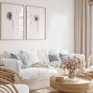 May include: A white couch with a throw blanket and pillows, a wooden coffee table with a vase of pink flowers, and two framed prints of dandelion flowers on the wall.