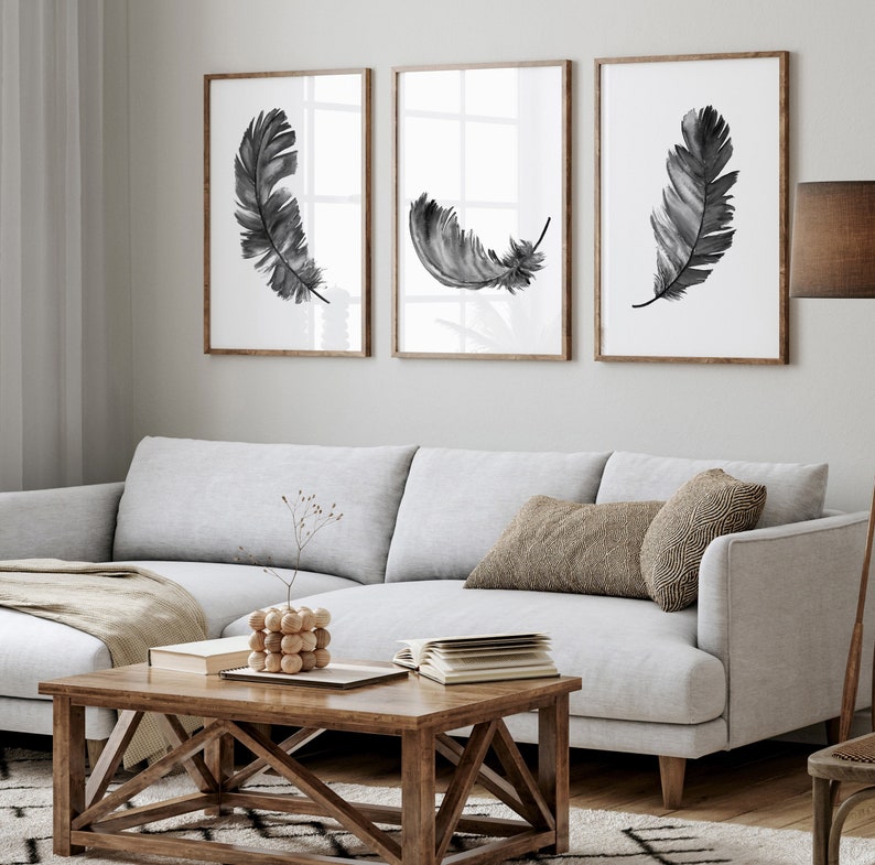 K&ouml;nnte beinhalten: Drei Schwarz-Wei&szlig;-Aquarell-Federdrucke in Holzrahmen, die an einer wei&szlig;en Wand &uuml;ber einem hellgrauen Ecksofa mit einem braunen Kissen und einem Holz-Couchtisch h&auml;ngen.