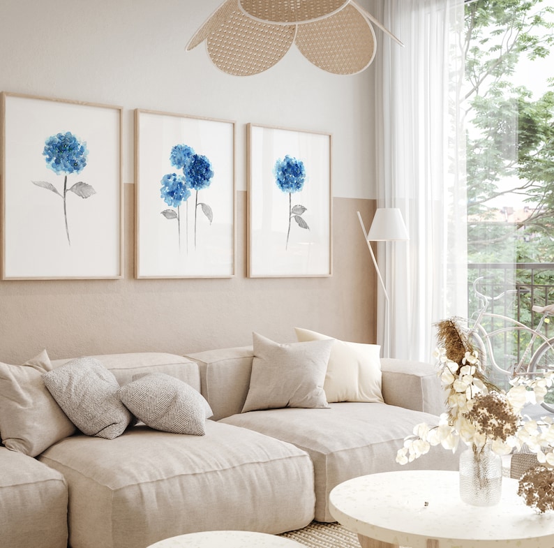 May include: Three framed watercolor paintings of blue hydrangeas on a white wall above a light beige couch with pillows. The couch is in a living room with a white coffee table and a vase of dried flowers.