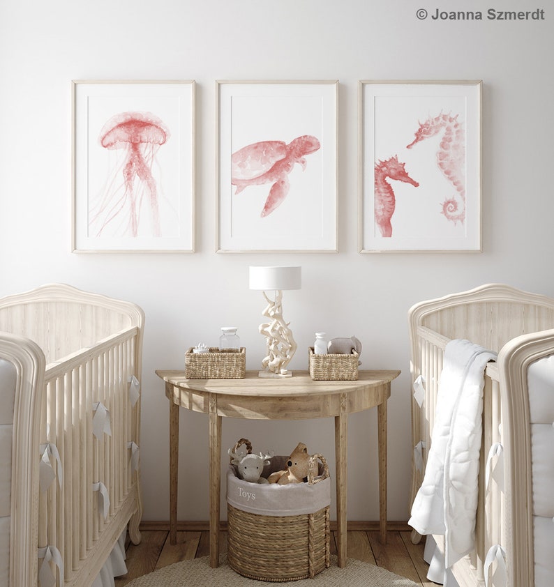 May include: Three watercolor prints of sea creatures in a nursery setting. The prints feature a jellyfish, a sea turtle, and a seahorse. The prints are in shades of pink and white and are framed in white.