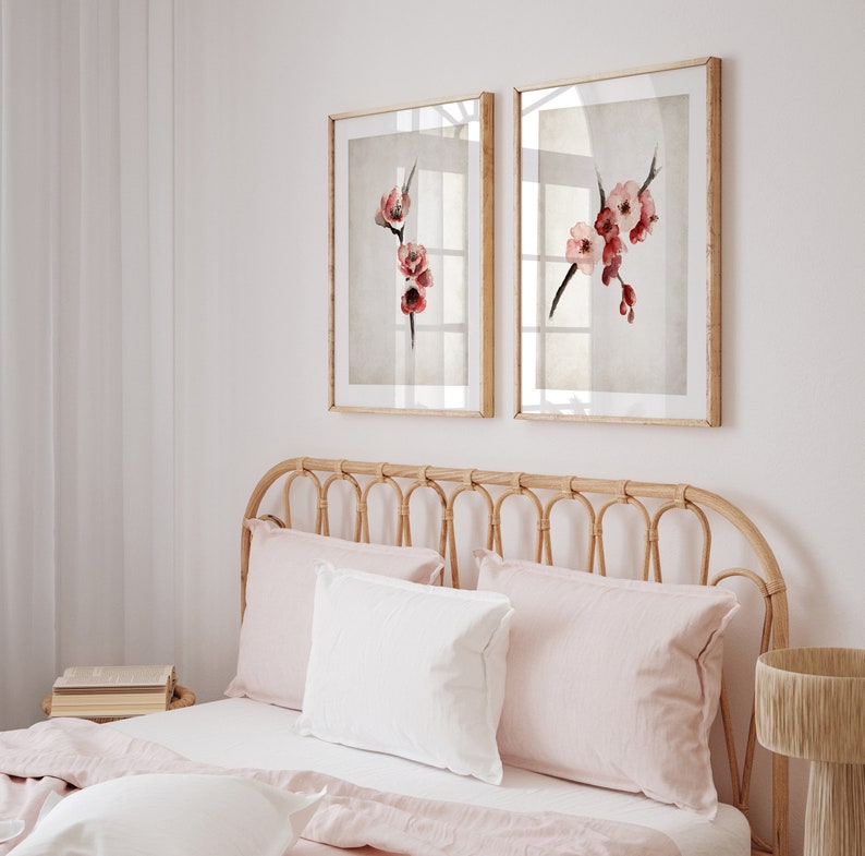 May include: Two framed watercolor paintings of pink and red flowers on a branch, hanging on a white wall above a light brown wicker headboard with two pink and white pillows.
