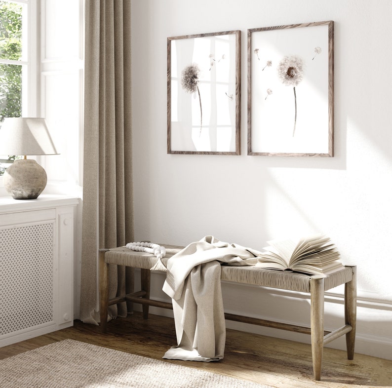 May include: A light wood bench with a woven seat and a white throw blanket draped over it. Two framed prints of dandelion flowers hang on the wall behind the bench.