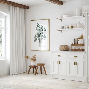 May include: A white kitchen cabinet with two drawers and two doors. The cabinet has gold hardware and a white countertop. A wicker basket and a small shelf with bottles and a ceramic jar are on the countertop. A framed watercolor print of eucalyptus leaves hangs on the wall above the cabinet.