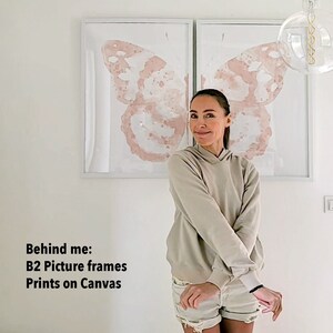 May include: Two large framed prints of a watercolor butterfly design on canvas. The prints are in a white frame and are hanging on a white wall. A person is standing in front of the prints.