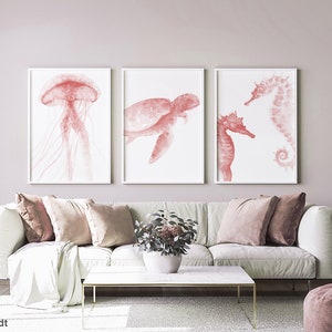 May include: Three watercolor prints of sea creatures in shades of pink. The prints feature a jellyfish, a sea turtle, and a seahorse. The prints are framed in white and are hanging on a light pink wall.