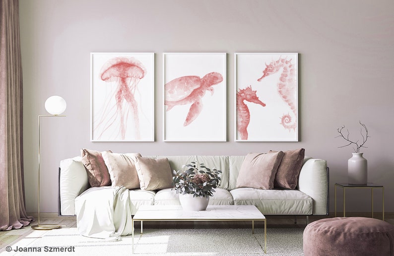 May include: Three watercolor prints of sea creatures in shades of pink. The prints feature a jellyfish, a sea turtle, and a seahorse. The prints are framed in white and are hanging on a light pink wall.