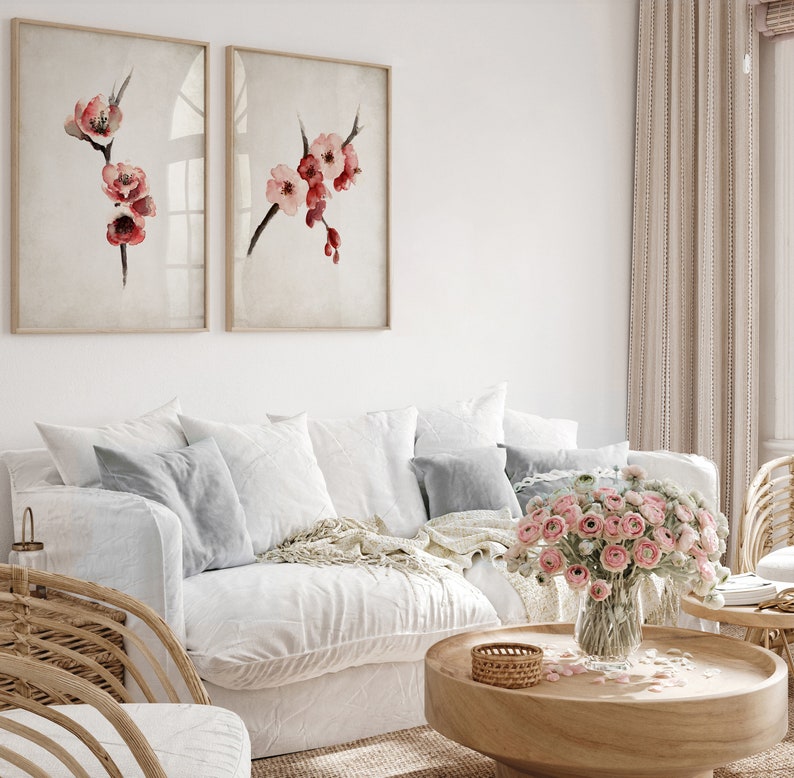 May include: A white couch with pillows and a throw blanket, a wooden coffee table with a vase of pink flowers, and two framed watercolor paintings of pink flowers on a white wall.