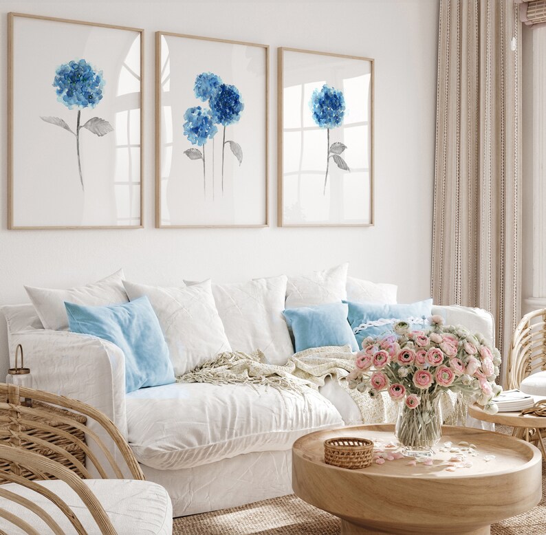 May include: A white couch with blue pillows and a throw blanket sits in a living room with three framed watercolor paintings of blue hydrangeas. A vase of pink flowers sits on a wooden coffee table.