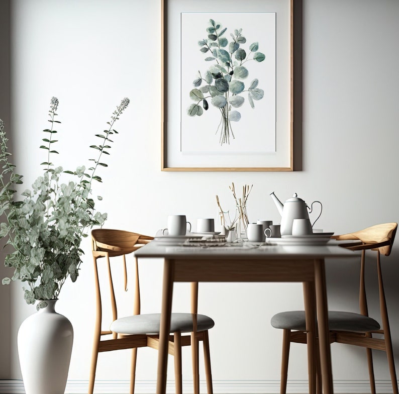 May include: A white ceramic vase with a leafy green plant sits on a wooden floor next to a dining table with four chairs. A framed watercolor print of a bouquet of eucalyptus leaves hangs on the wall behind the table.