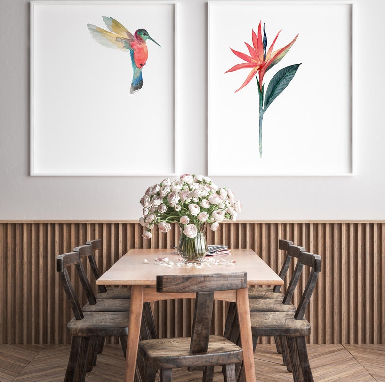 May include: A dining room table setting with a bouquet of white flowers in a glass vase, two framed watercolor prints of a hummingbird and a bird of paradise flower, and six wooden chairs.