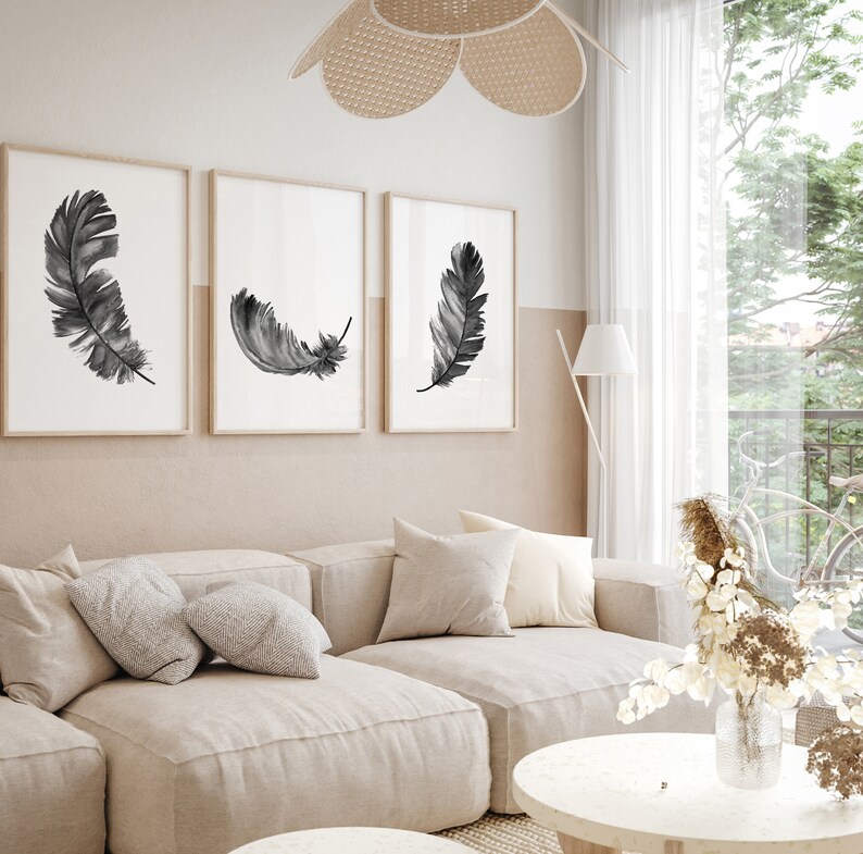K&ouml;nnte beinhalten: Drei gerahmte Schwarzwei&szlig;-Aquarell-Federdrucke an einer beigefarbenen Wand &uuml;ber einem hellbeigen Ecksofa mit Kissen. Eine Vase mit Trockenblumen steht auf einem runden wei&szlig;en Couchtisch.