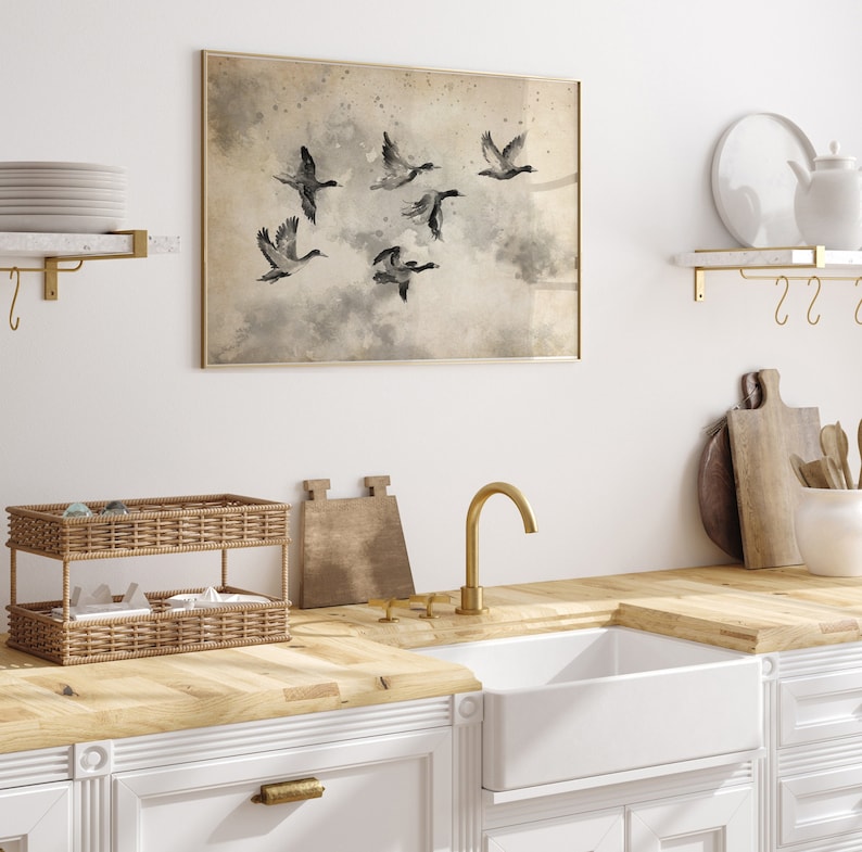 May include: A framed watercolor print of six ducks in flight, set against a muted, cloudy background. The artwork is displayed above a kitchen sink with a gold faucet, white cabinets, and a woven basket.