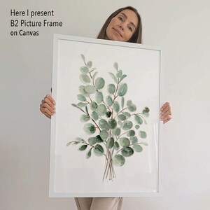 May include: A white picture frame with a watercolor painting of a bouquet of green eucalyptus leaves on a white background. The frame is being held by a person.