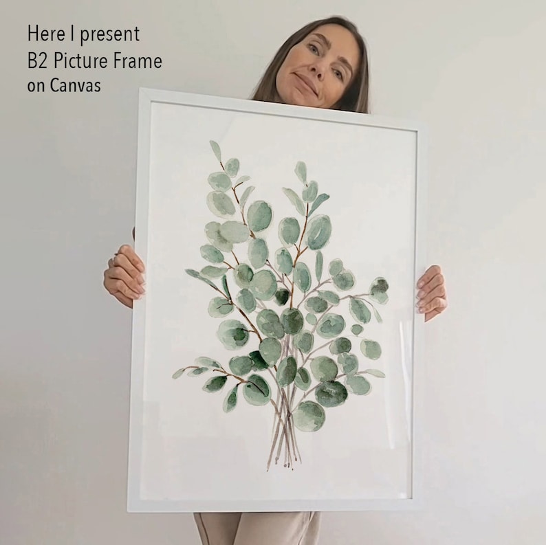 May include: A white picture frame with a watercolor painting of a bouquet of green eucalyptus leaves on a white background. The frame is being held by a person.