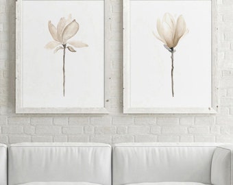 Könnte beinhalten: Zwei gerahmte Aquarellbilder von Magnolienblüten in Beige- und Weißtönen vor einem Hintergrund aus weißen Backsteinwänden.